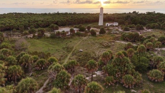 Aerial view of a white lighthouse among palm trees at sunset, with the sea beyond